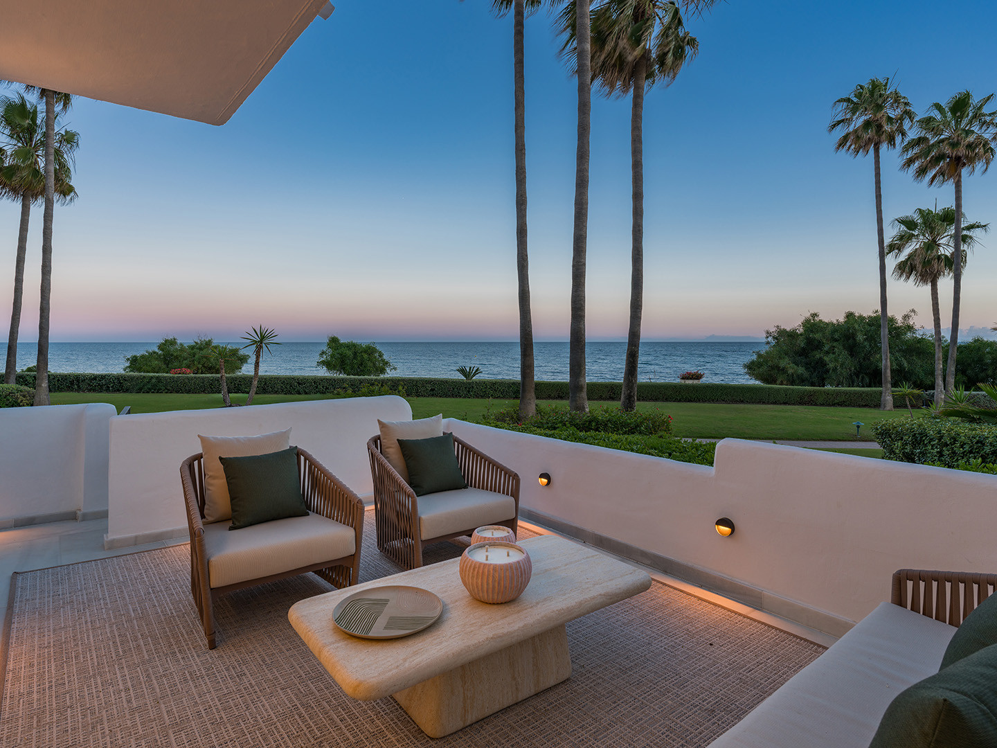 Patio with outdoor furniture at dusk, facing the ocean and palm trees, with warm wall lighting.