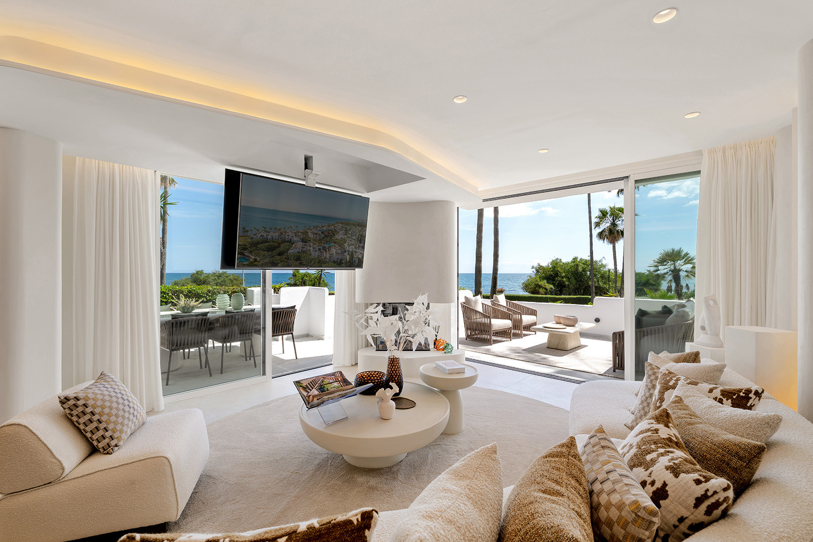 Bright living room with large TV, modern fireplace, and ocean views through expansive sliding glass doors.