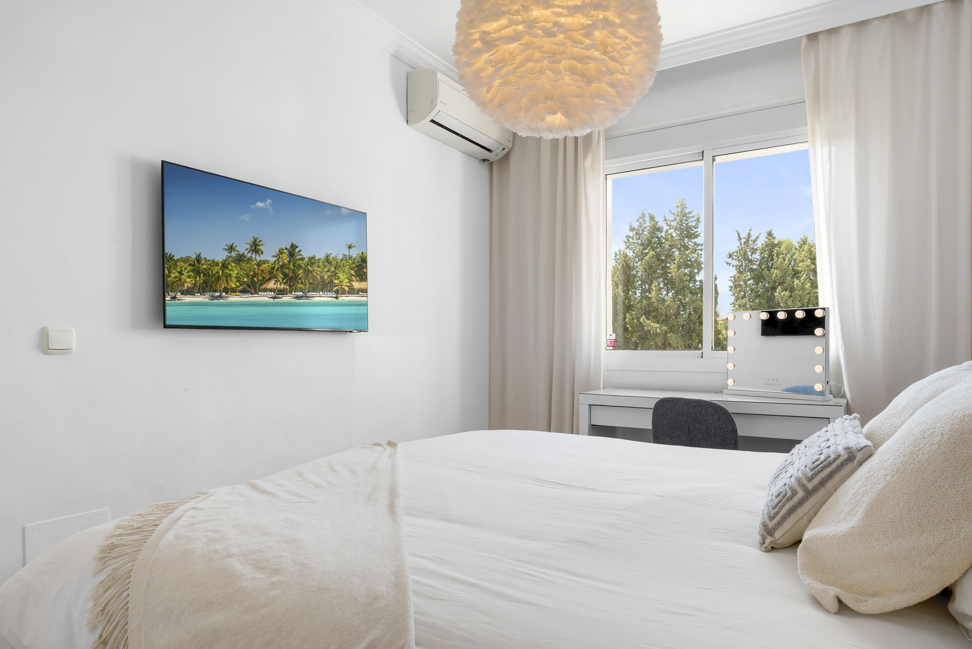 Bedroom featuring a gray headboard, table lamp, and a mounted TV facing a window with dense trees outside.