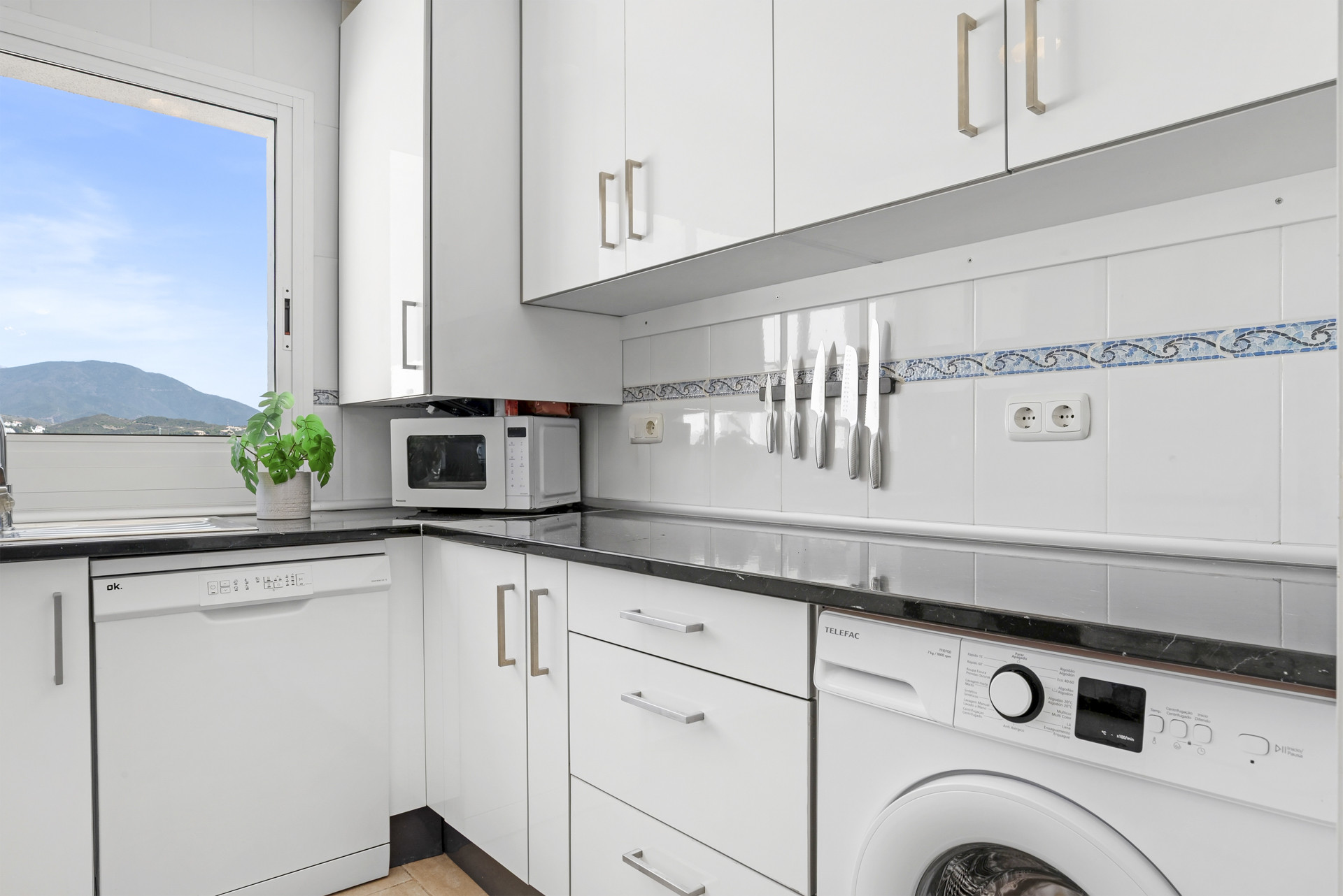 Modern kitchen with white cabinets, black countertops, and mountain view through large window.