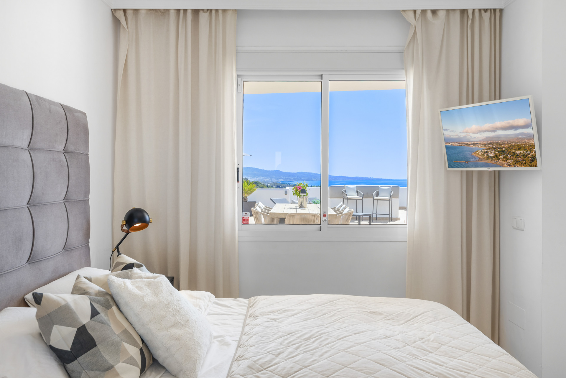 Bedroom with gray headboard and curtains, showcasing a distant view of the sea through large windows.
