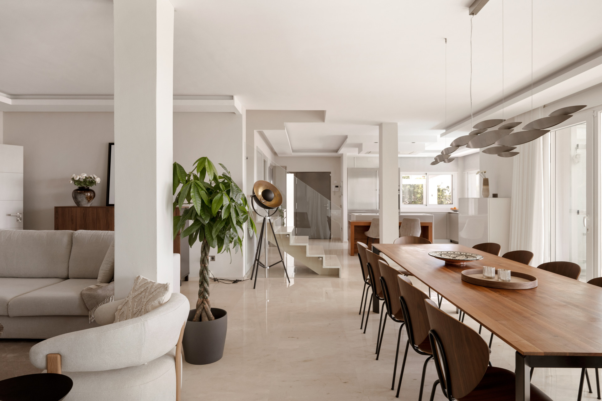Dining area with a long wooden table, surrounded by chairs, leading to the kitchen area.