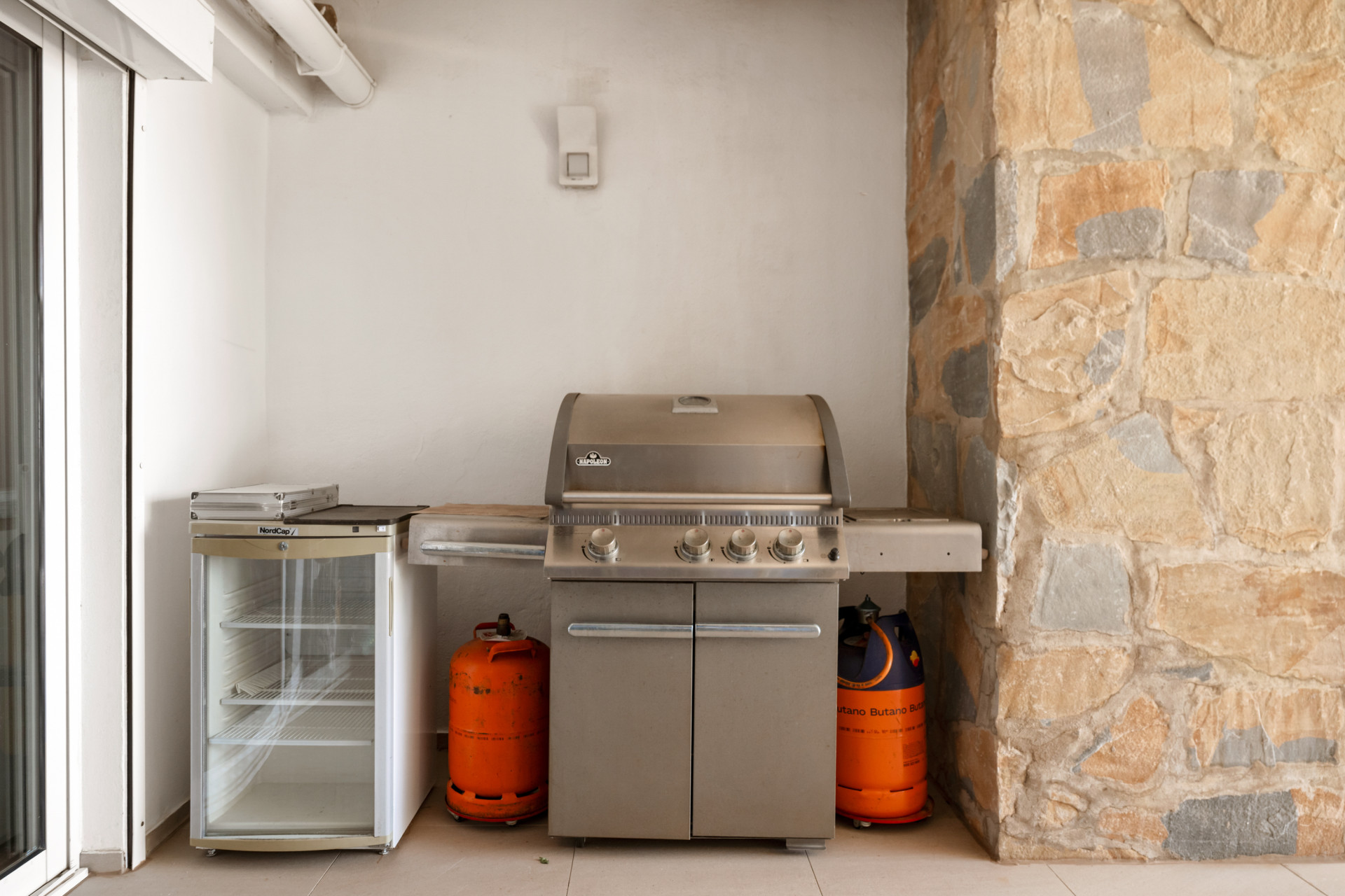 Outdoor kitchen area with a built-in grill and gas tanks beside a glass door.