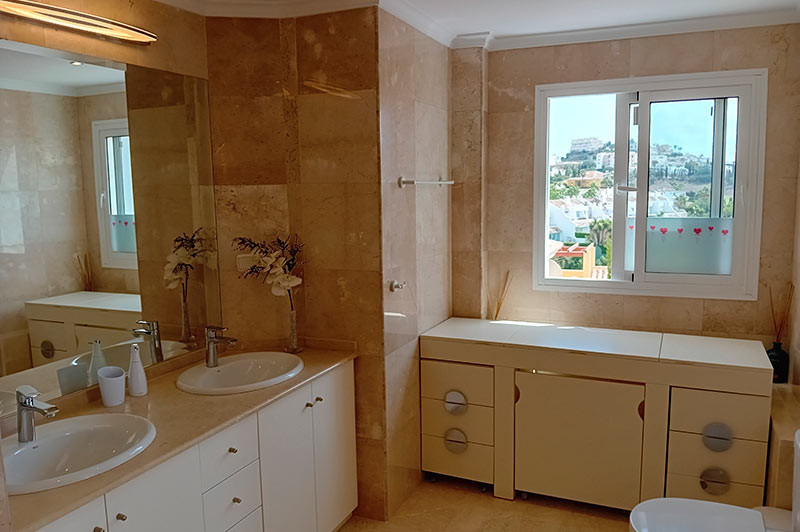 Bathroom with double sinks, a large mirror, beige tiled walls, and a window overlooking a town.