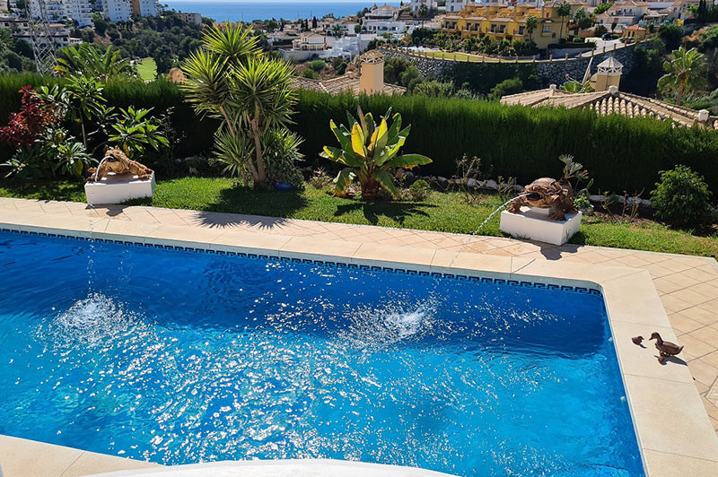 Outdoor swimming pool with bubbling fountains, surrounded by green plants and distant houses and sea.