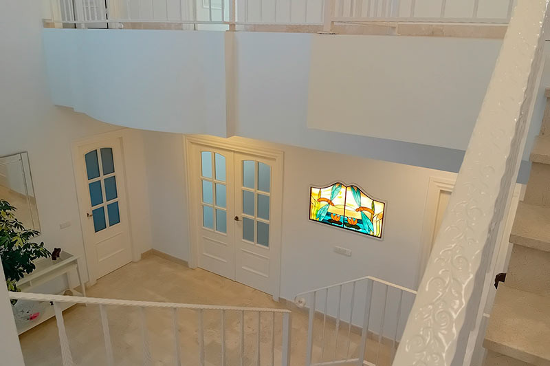 Staircase view of a bright hallway with white doors, light-colored flooring, and stained glass art.
