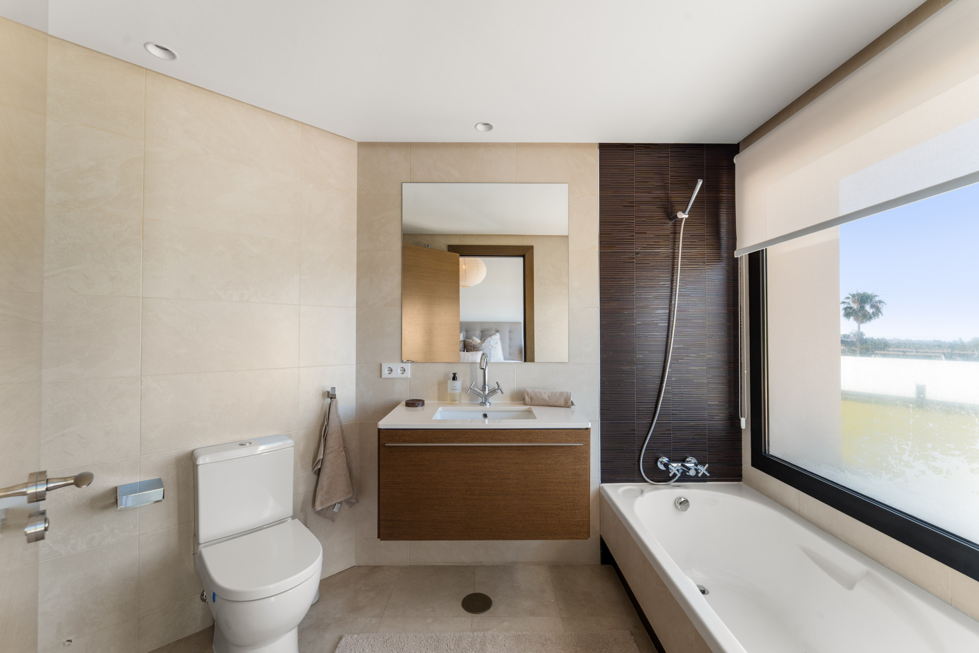Modern bathroom with beige tiles, large mirror, and bathtub beside a window.