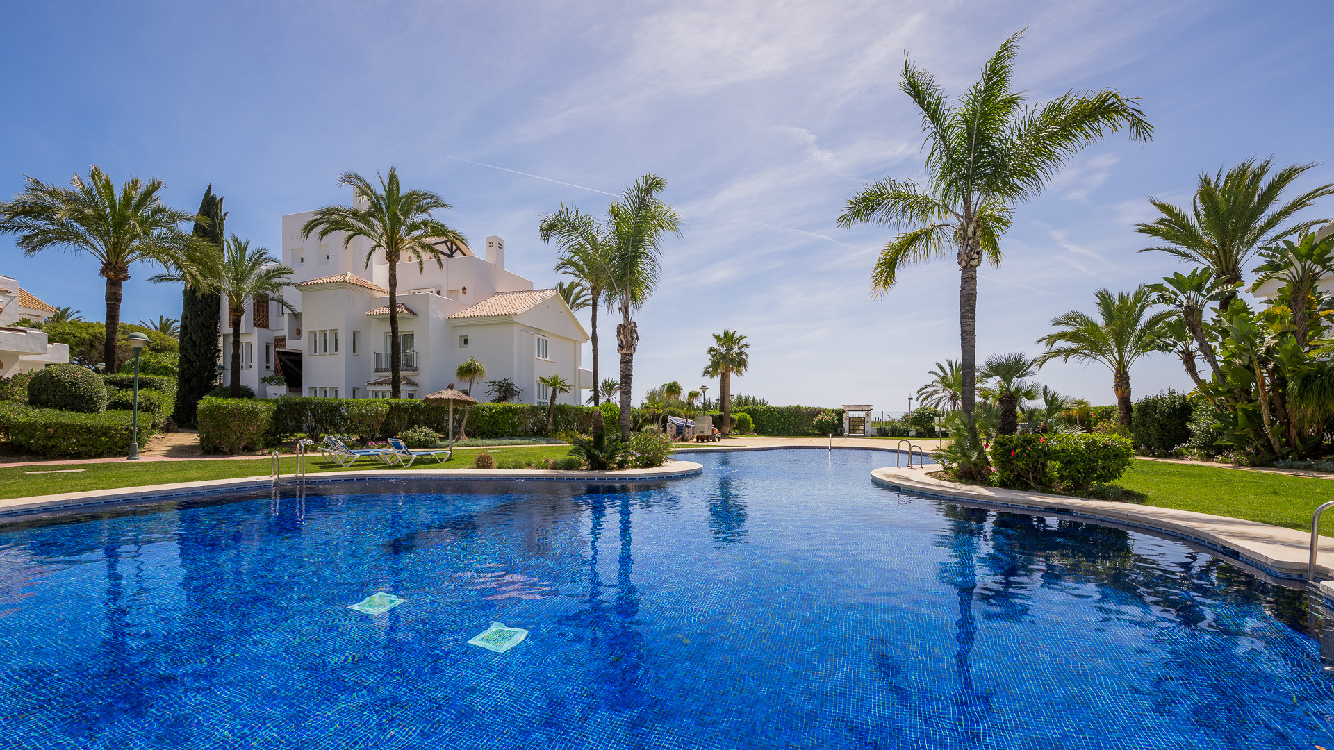 Large blue tiled swimming pool with palm trees, green lawns, and white buildings under a sunny sky.