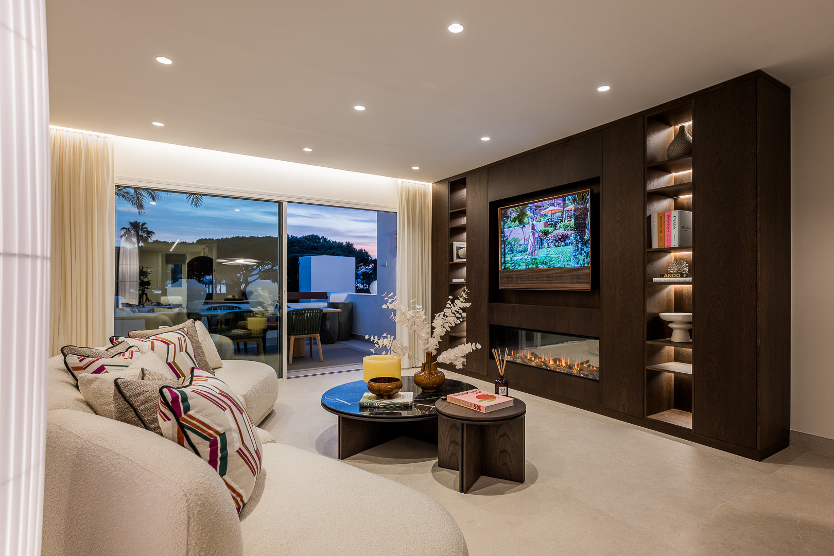 Living room with white curved sofa, coffee tables, television over fireplace, and glass doors to terrace.