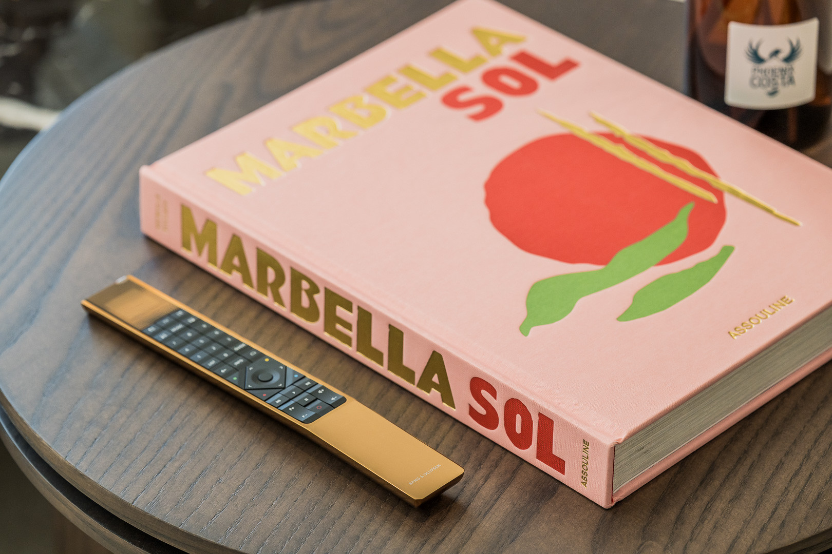 A gold remote control and a "Marbella Sol" book rest on a dark wood table.