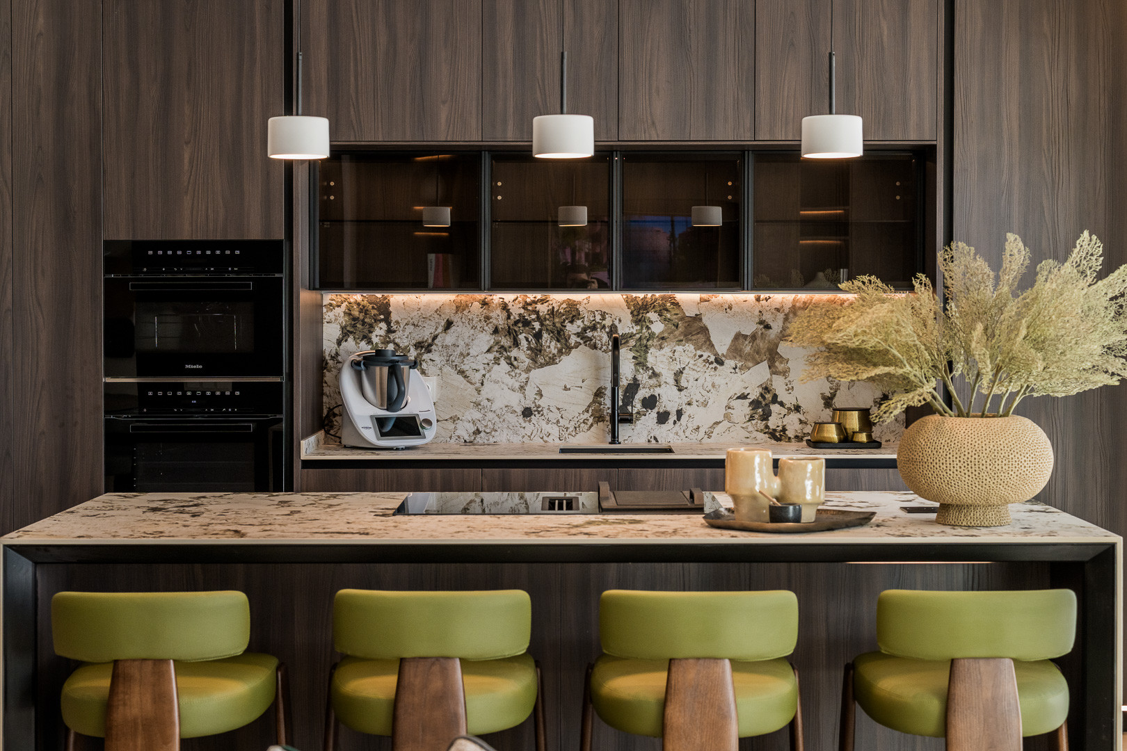 Modern kitchen with wood cabinets, stone island with green stools, built-in ovens, and pendant lights.