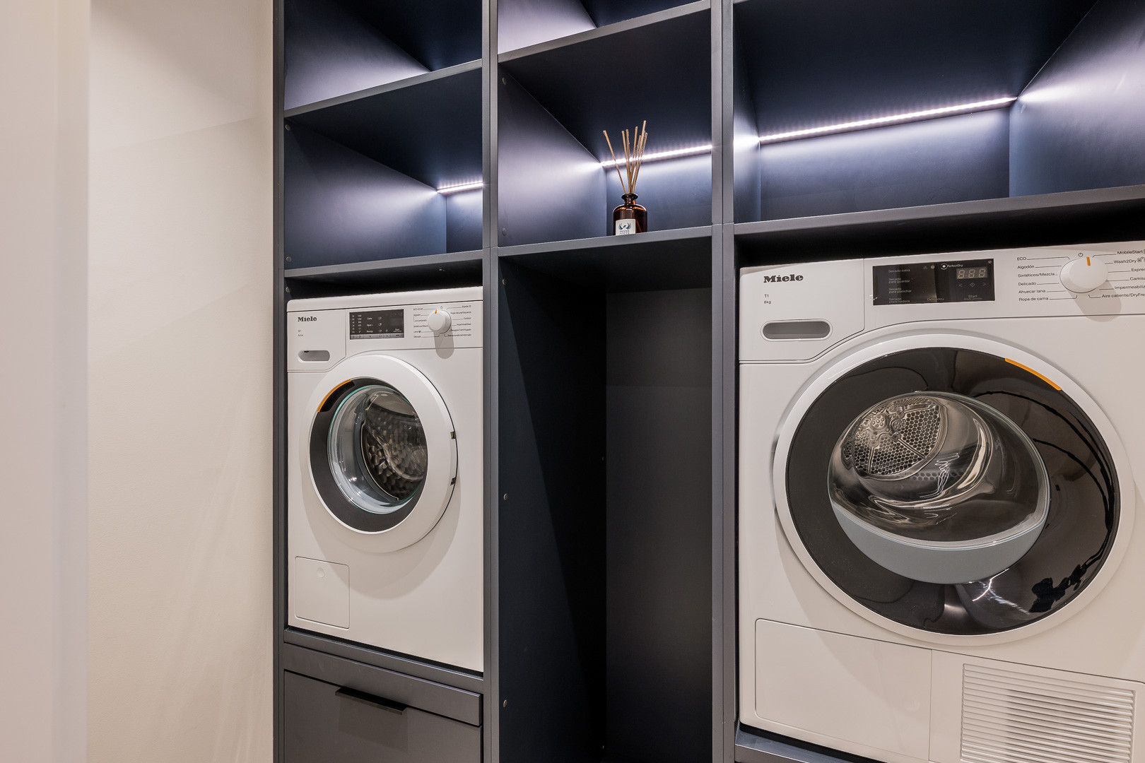Laundry area with two white Miele washer machines integrated into dark grey shelving with lighting.