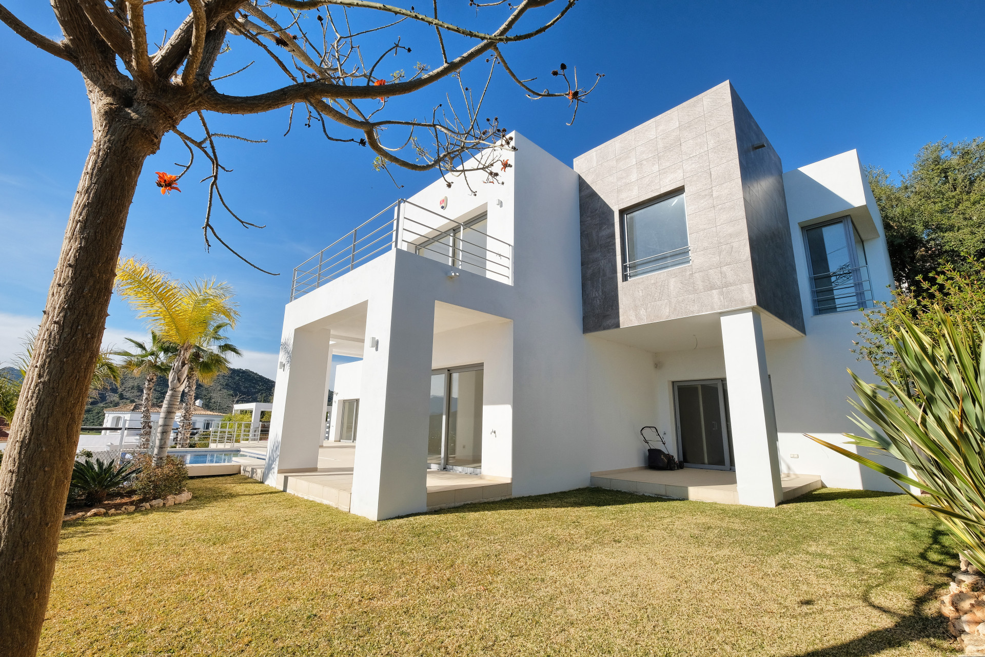 Exterior shot of a two story modern villa with white and grey facade, green lawn, and sparse trees.