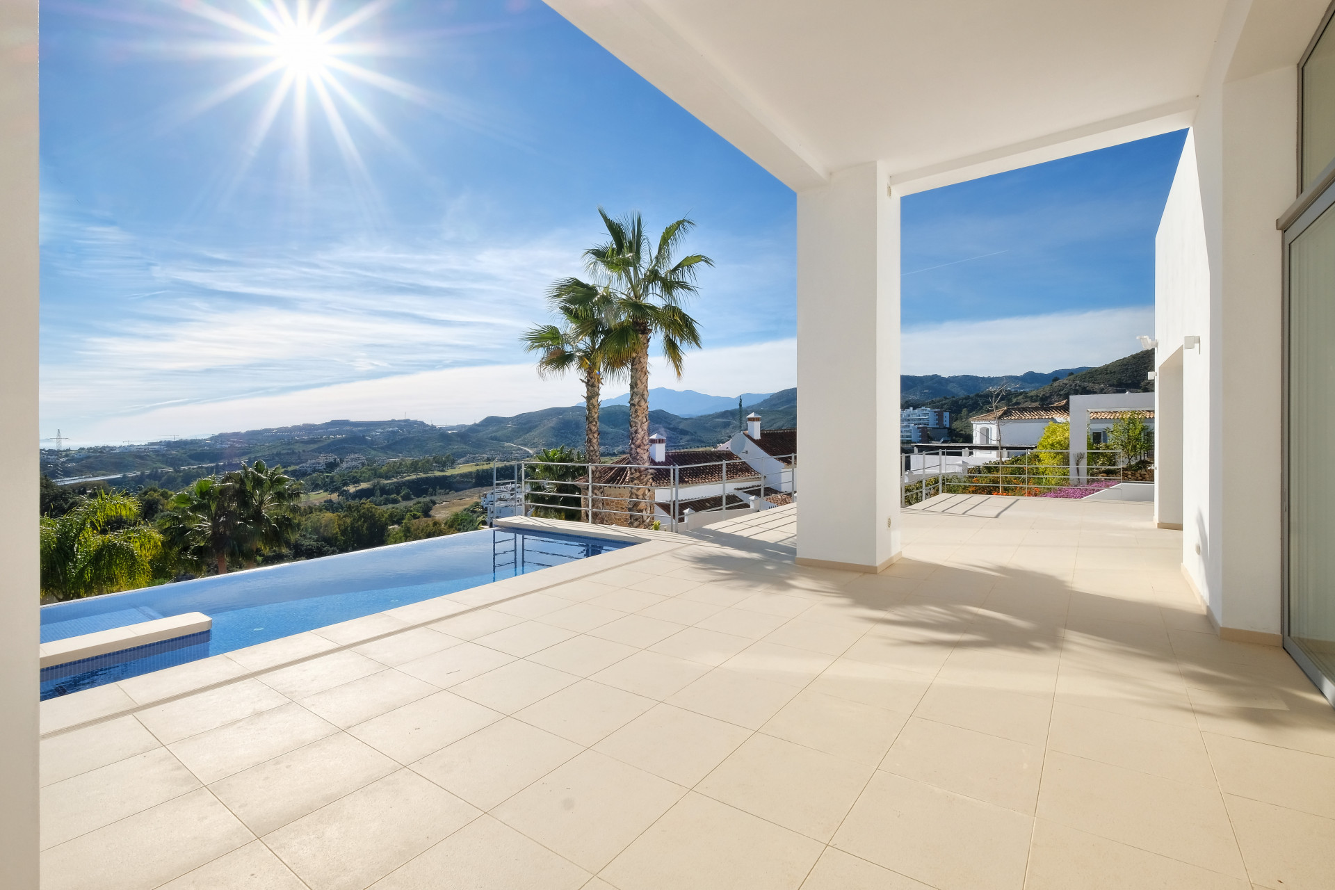 Covered patio overlooking an infinity pool, mountains, and sunlit sky.