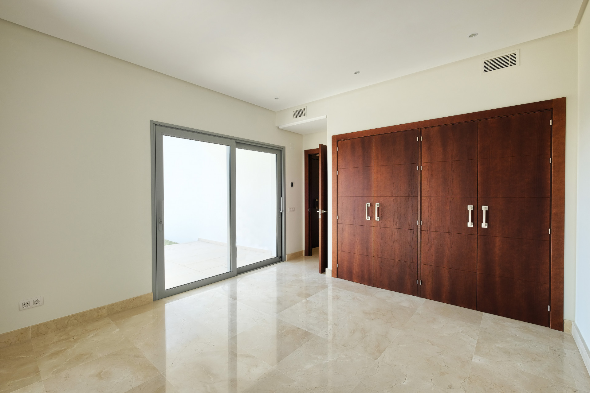 Bedroom with glossy marble floor, white walls, large sliding glass doors, and dark wood built-in wardrobes.