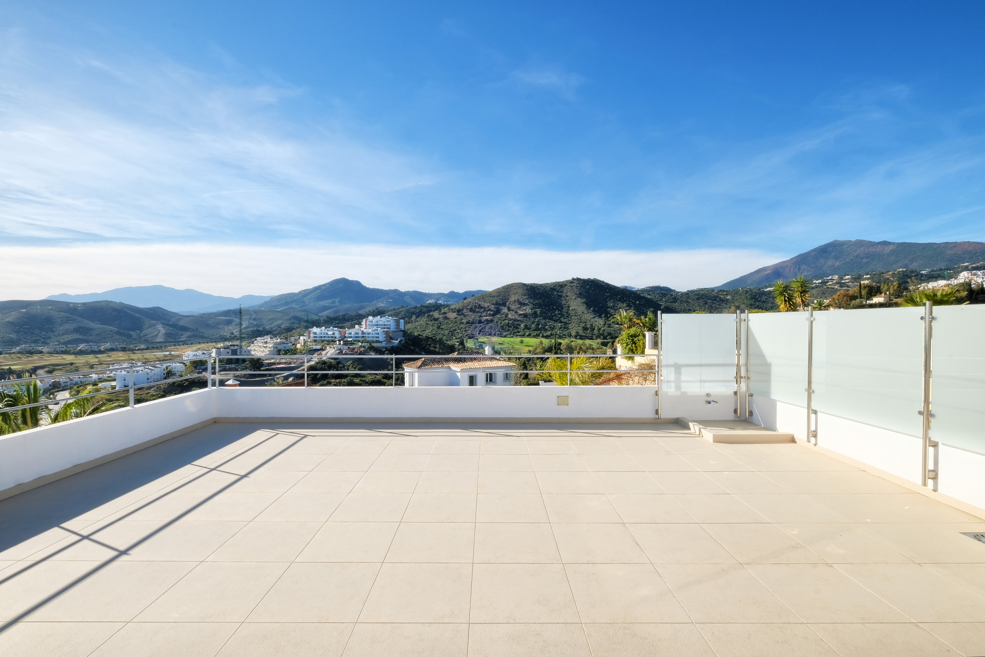 Rooftop terrace with beige tiles and glass railings overlooking a mountainous village under a blue sky.