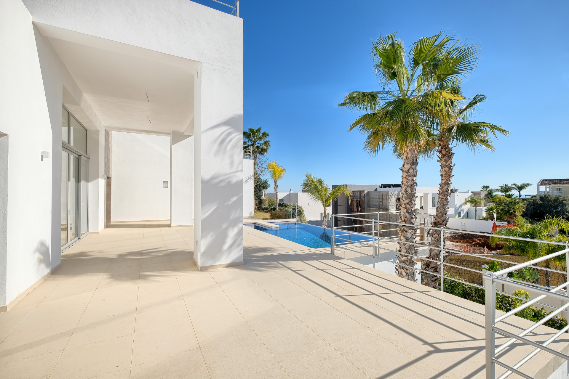 Outdoor terrace with beige tiles and metal railings, overlooking a swimming pool and distant palm trees.