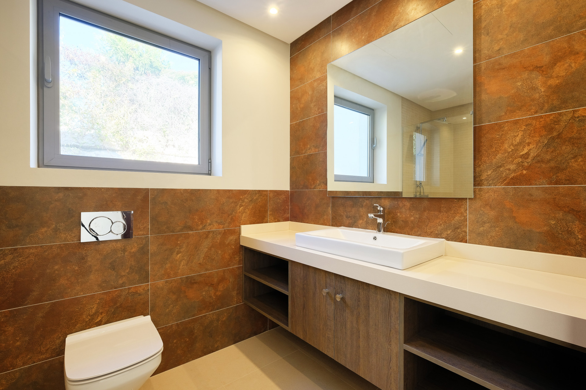 Bathroom with rectangular mirror, white sink, wooden vanity, and brown tiled walls. Window shows greenery.