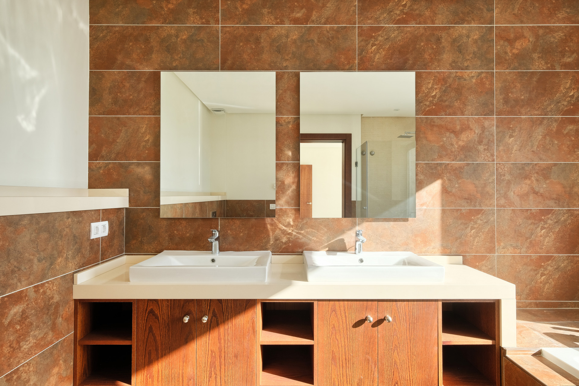 Bathroom vanity with two white sinks, light countertop, and wooden cabinets, against a brown tiled wall.