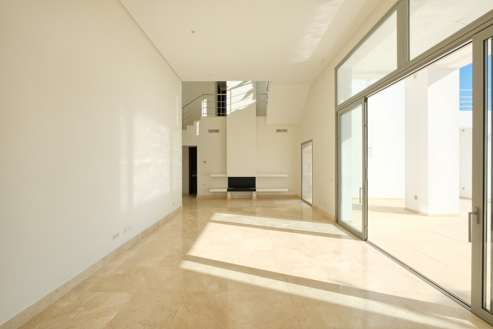 Empty living space with marble floors, a large sliding glass door, and a floating staircase in the background.