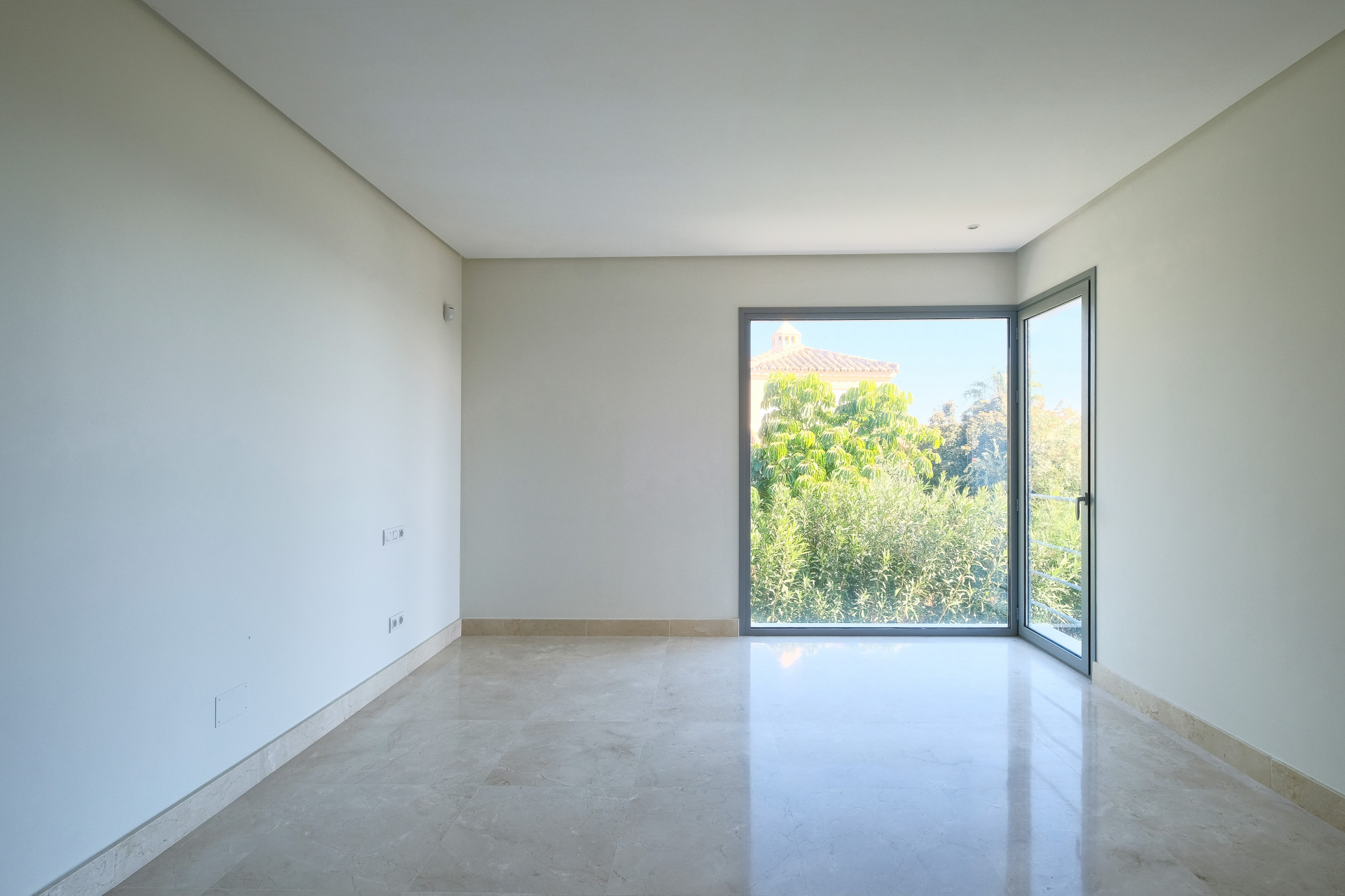 Empty room with light-colored marble floor and large glass door opening to outdoor greenery and a house.