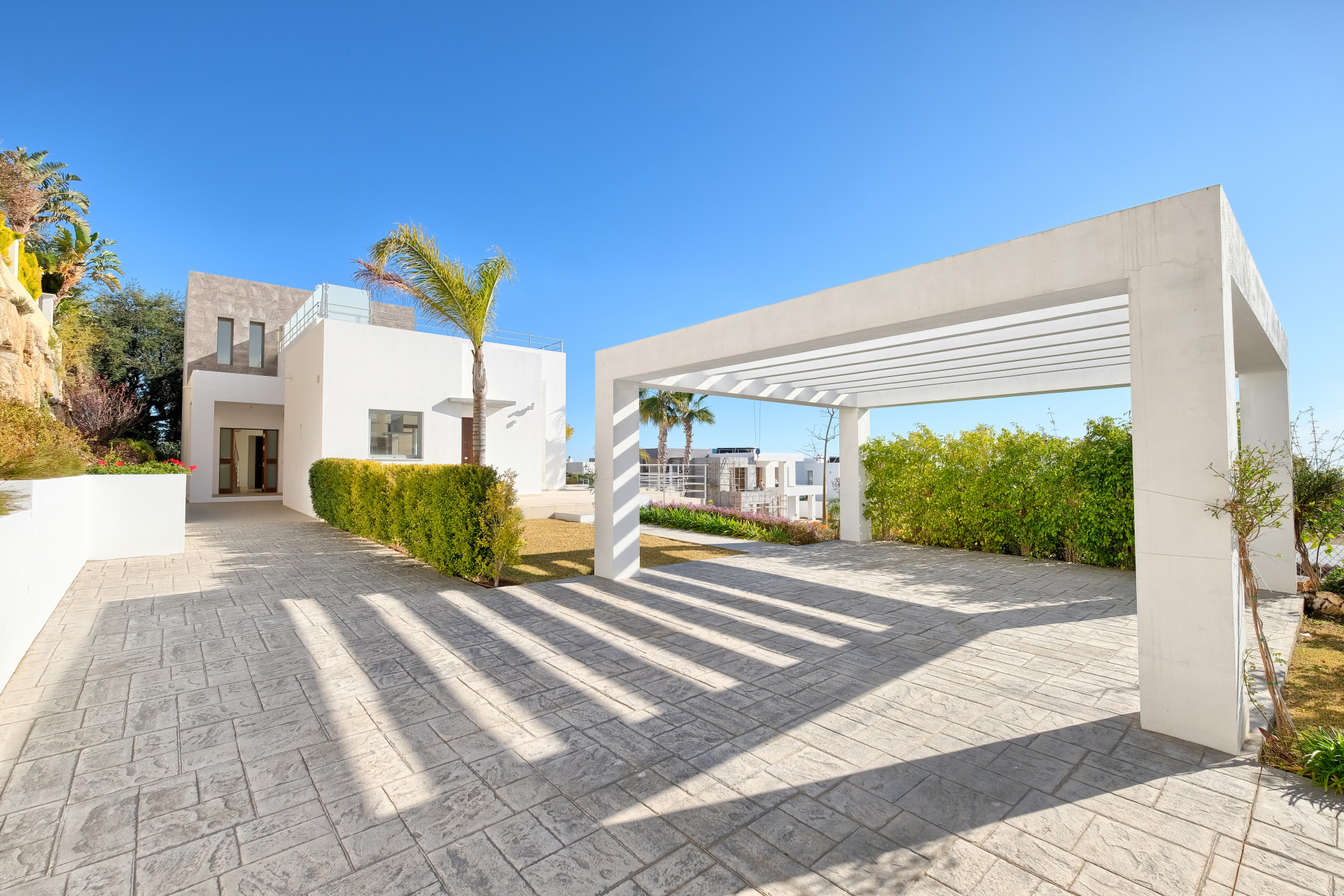 View of a modern white villa and carport from the patterned driveway, with palm trees and blue sky.