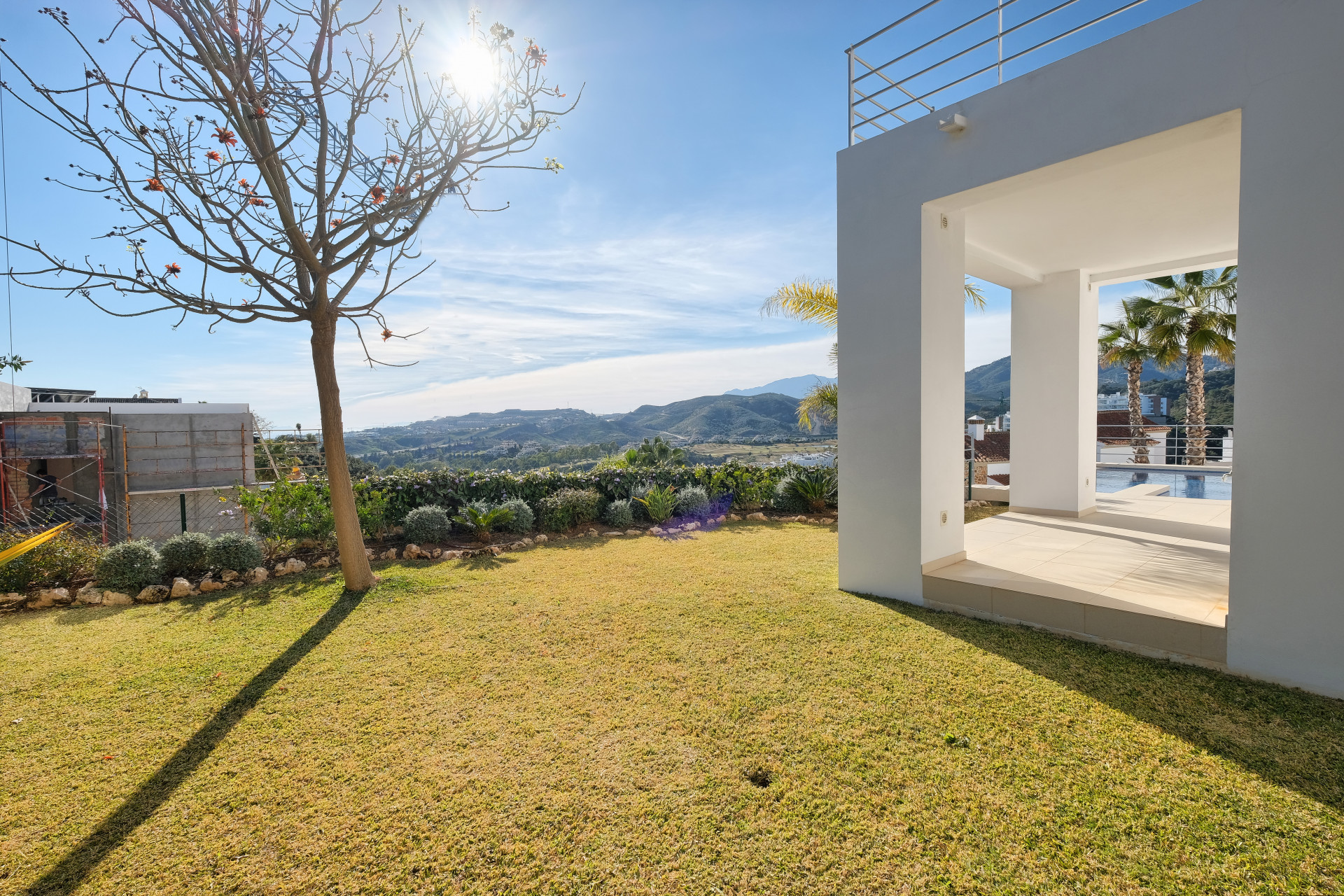 View from a green lawn towards a white villa terrace, with mountains, sky, and pool visible in the distance.