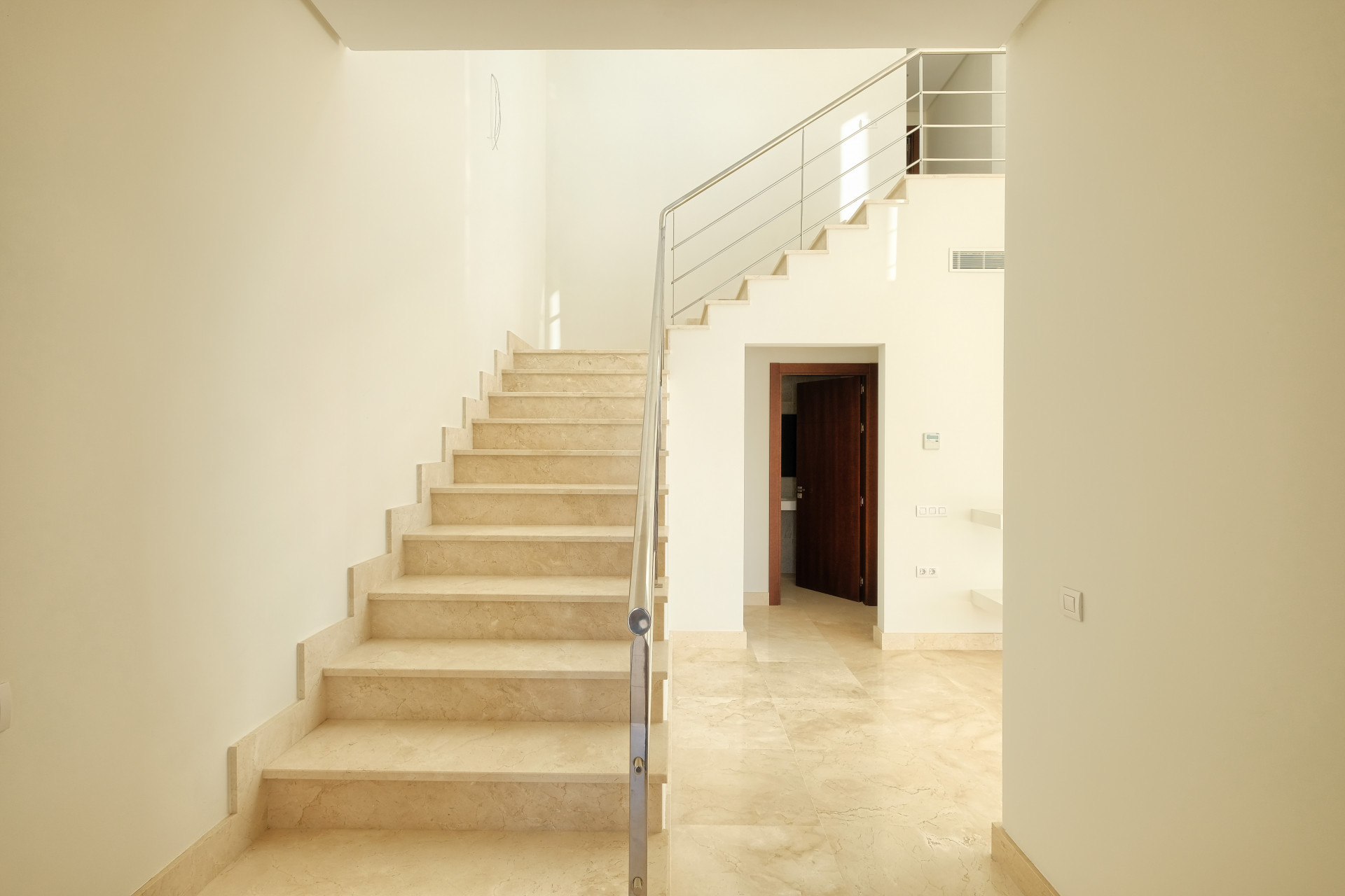 View of a spacious, light-colored marble staircase with a metal handrail. An open wooden door is visible below.
