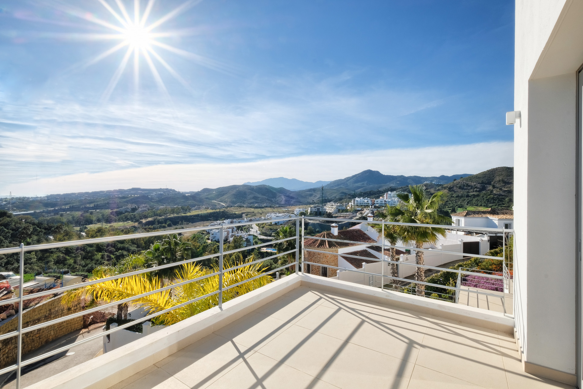 Outdoor balcony with tile floor and metal railing, overlooking mountains, town, and palm trees under a bright sun.