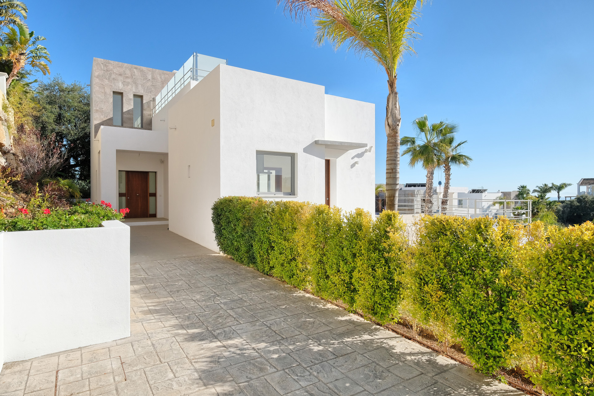 Front exterior of a white modern villa with a wooden door, surrounded by hedges and a paved driveway.