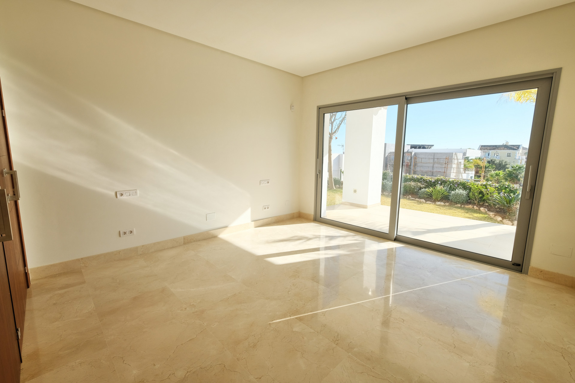 Empty room with light marble flooring and a sliding glass door leading to a garden area. Sunlight streams in.