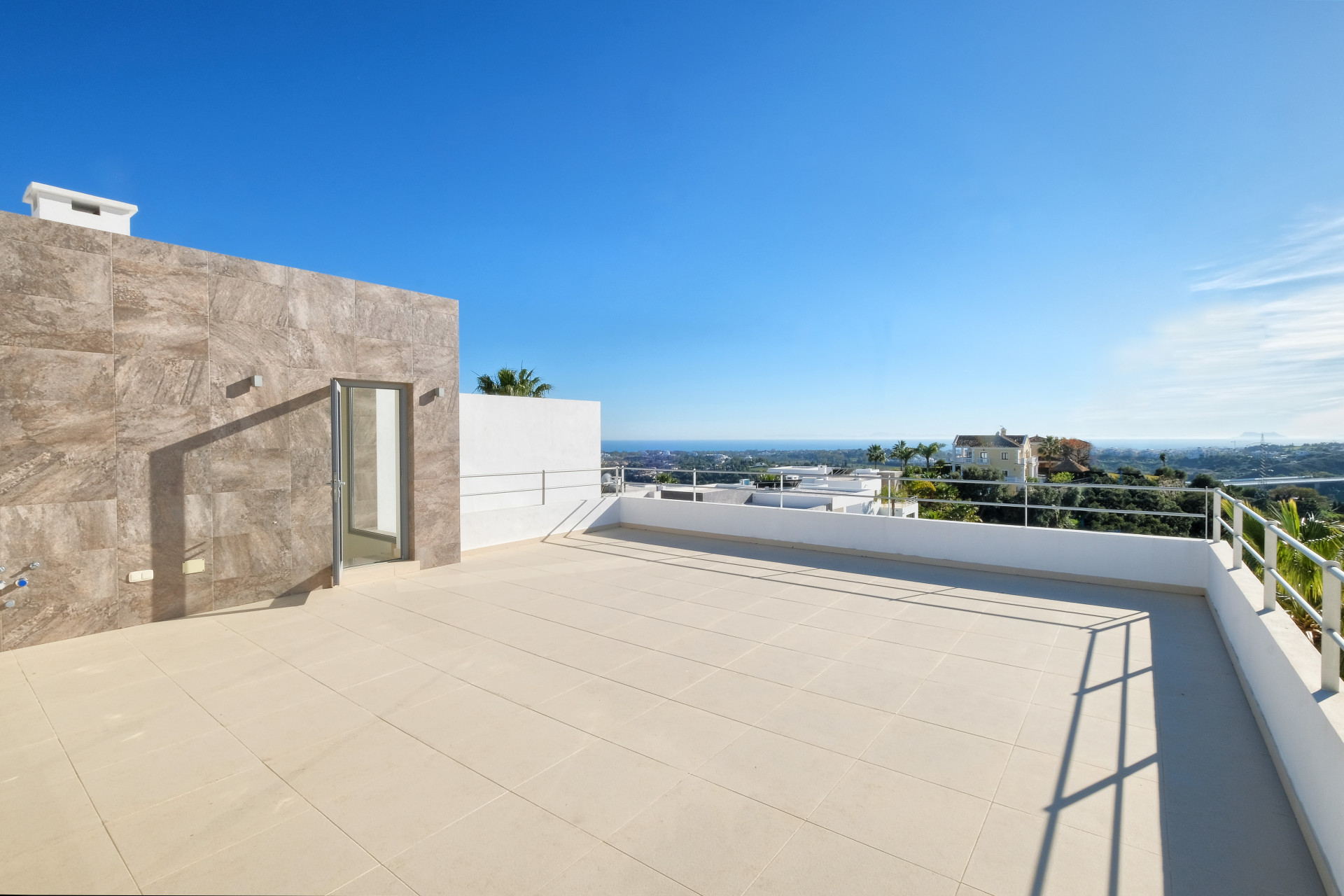 Rooftop terrace with tiled floor, white railing, a door on a tiled wall, and panoramic sea and mountain views.