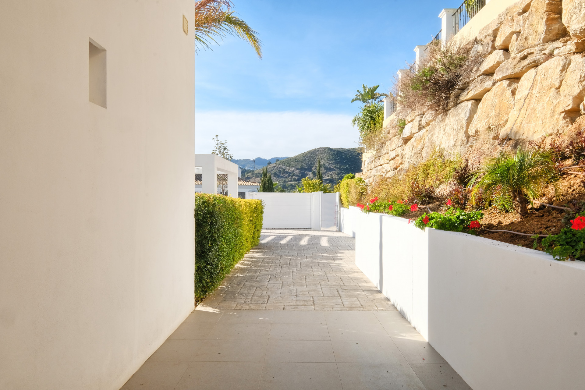 Exterior pathway with white walls and hedges, leading to a gate with a mountainous backdrop.