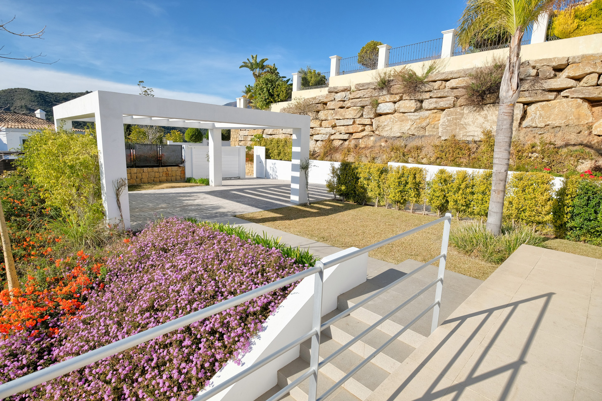 Exterior view of a white pergola structure over a lawn, with flowering plants and stone walls.