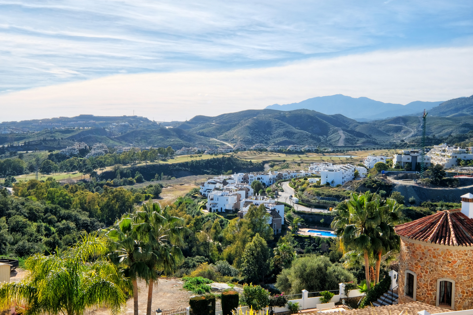 Panoramic view of a hillside village with white buildings, palm trees, and distant mountains.