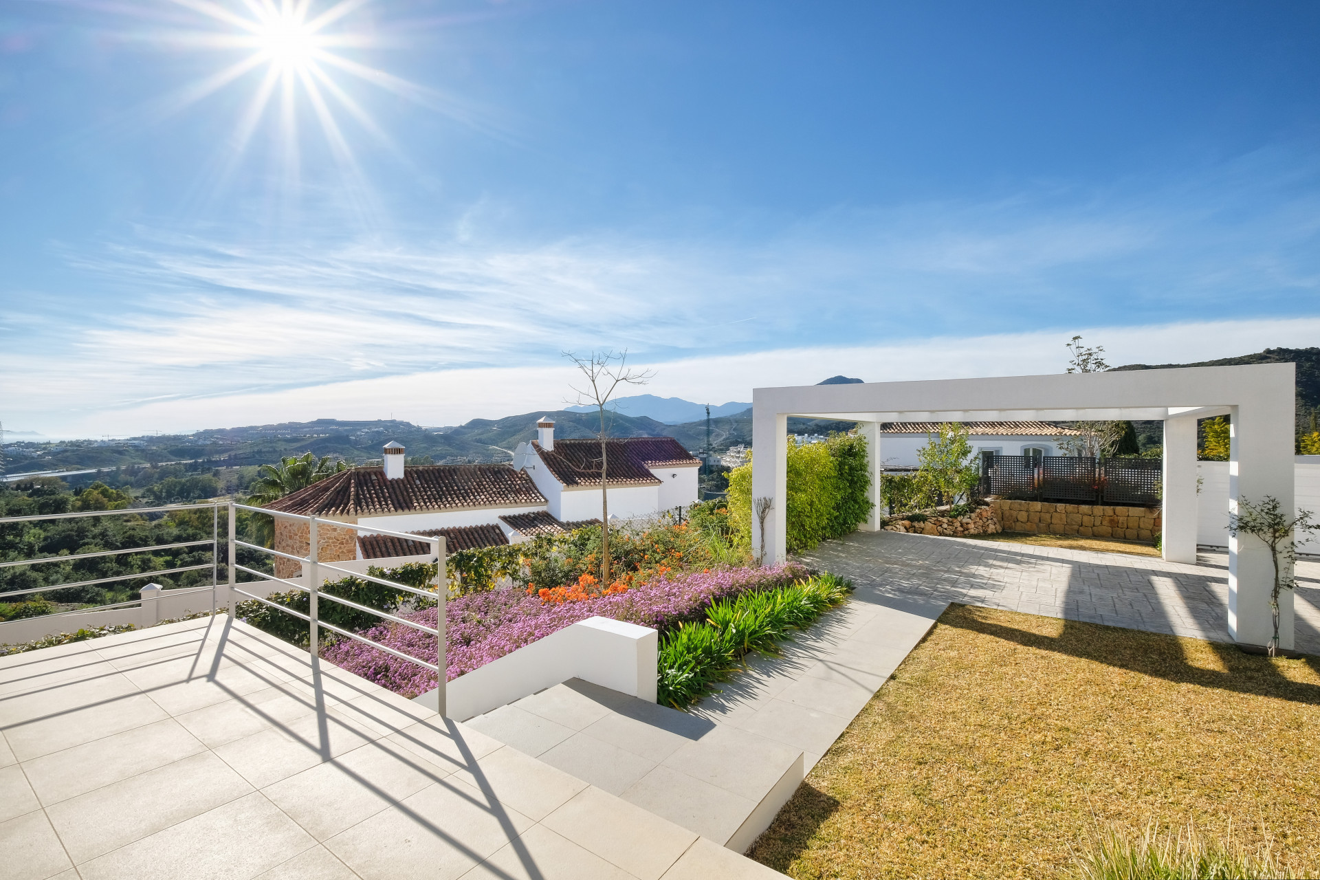 Outdoor tiled patio with white railings, leading to a lawn, a white pergola, and distant mountains.