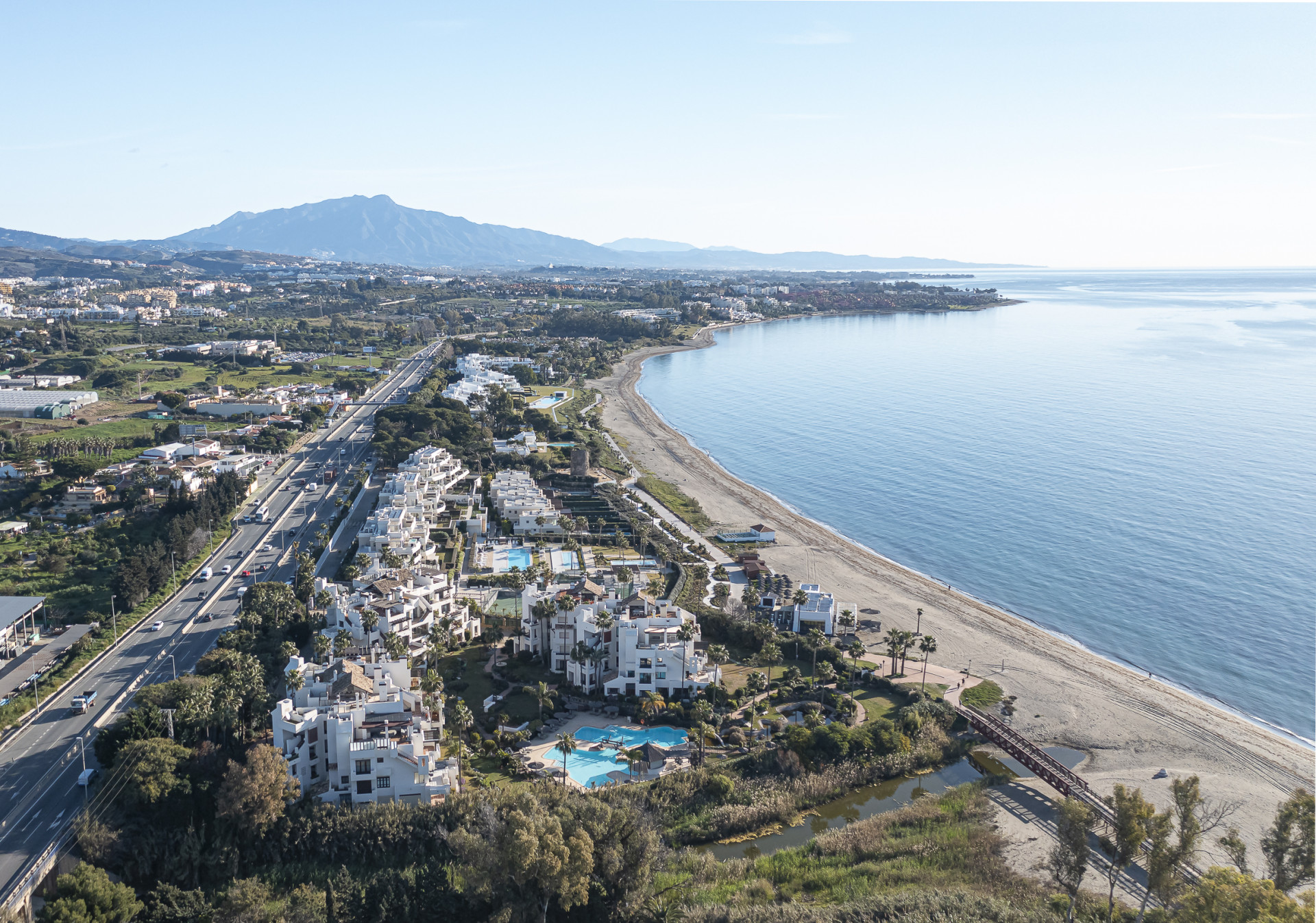 Aerial view of coastal development along a beach, with a road and mountains in the distance.