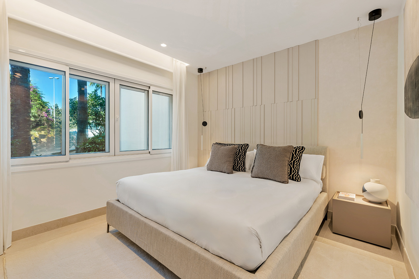 Bedroom with large window overlooking greenery, a bed with light bedding, and a textured headboard.