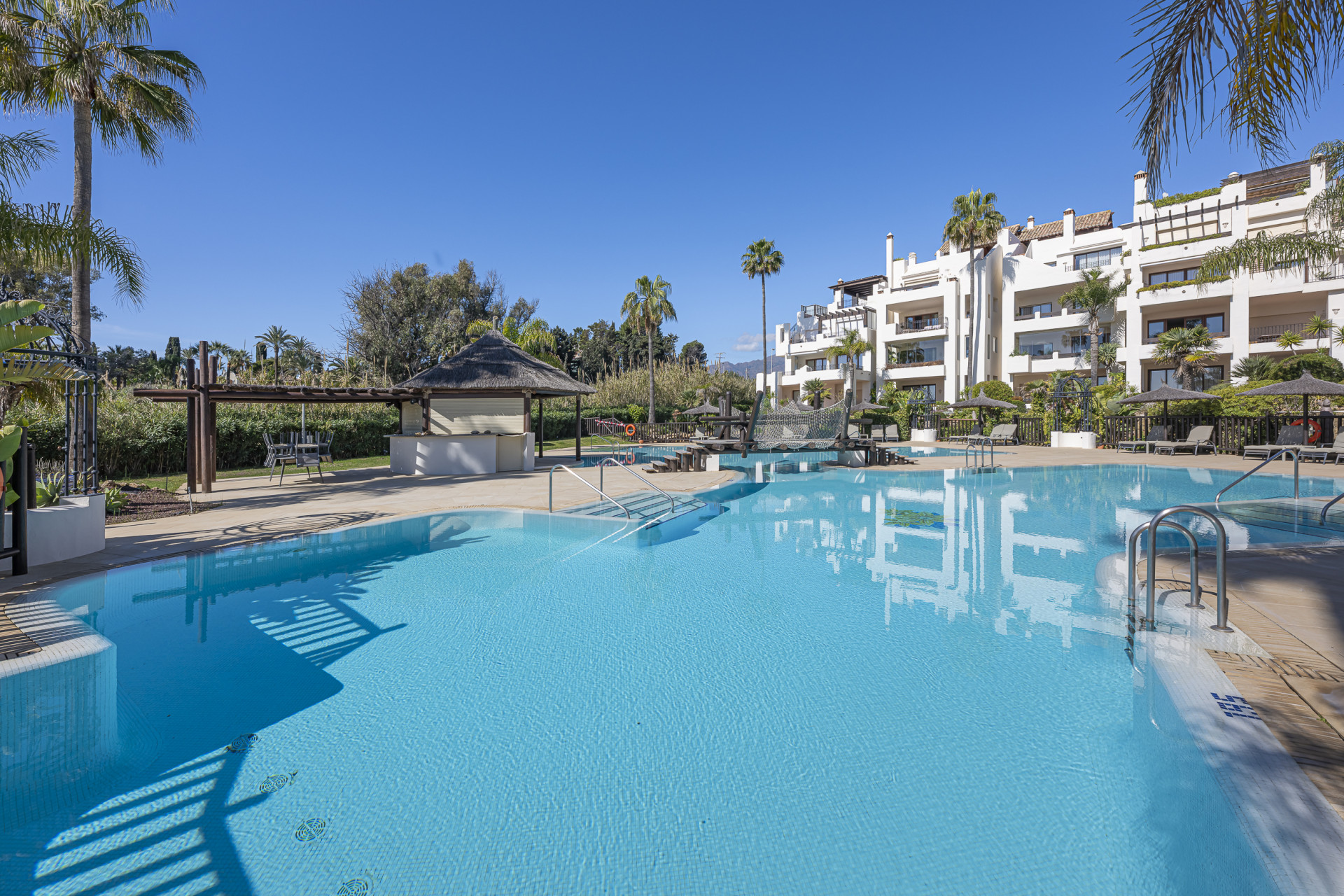 Expansive outdoor swimming pool with a thatched-roof cabana and apartment complex in the background.