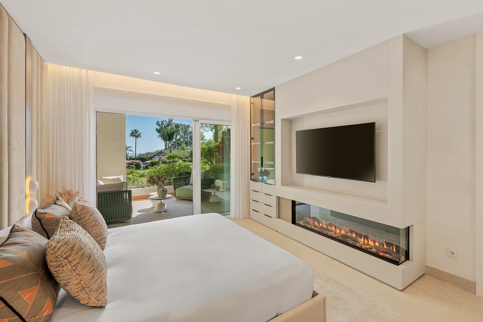 Bedroom with a large bed, wall-mounted television, and a fireplace. Sliding doors open to a patio and garden.