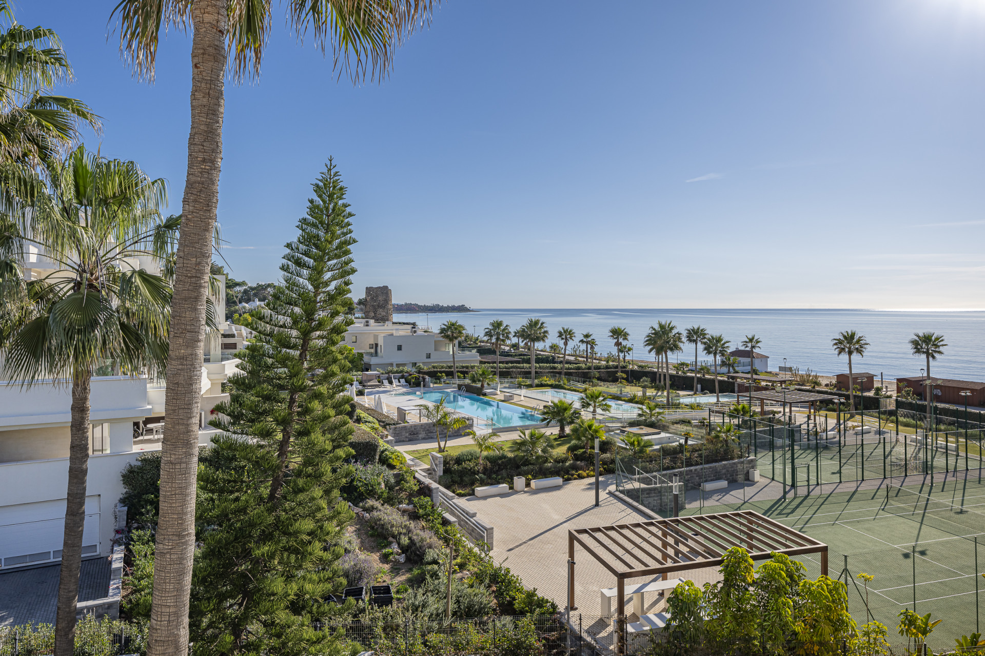 Overhead view of resort buildings, multiple pools, tennis courts, and the ocean under clear skies.