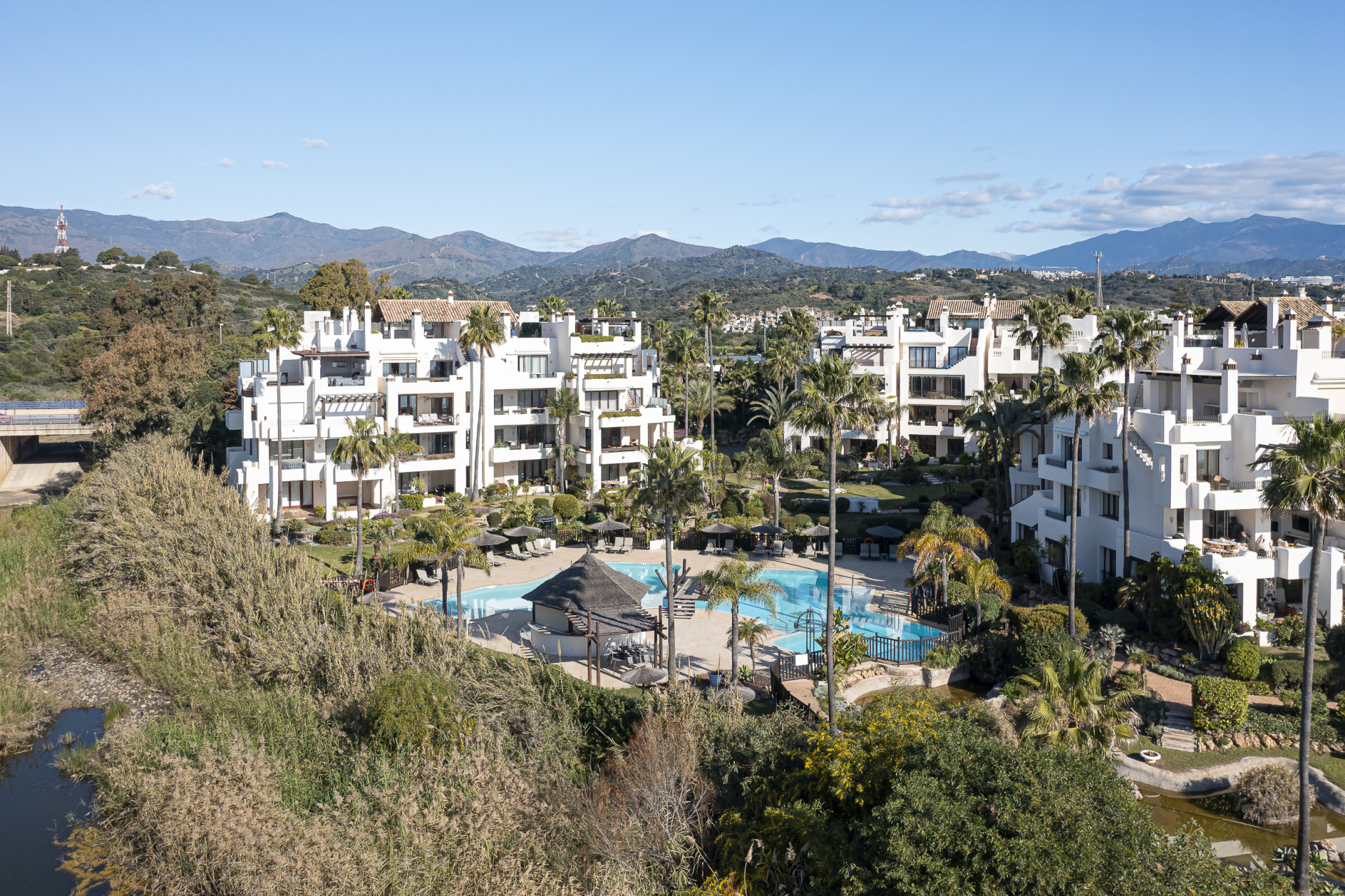 Aerial view of white apartment buildings with pools, palm trees, and lush landscaping, backed by mountains.