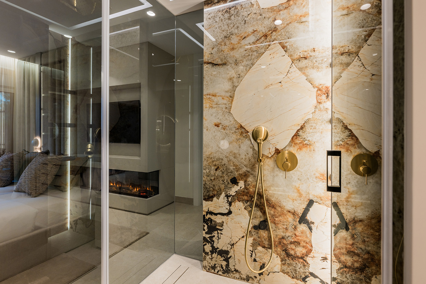 Detail of a glass shower with gold fixtures and marble walls. The bedroom and fireplace are visible through the glass.