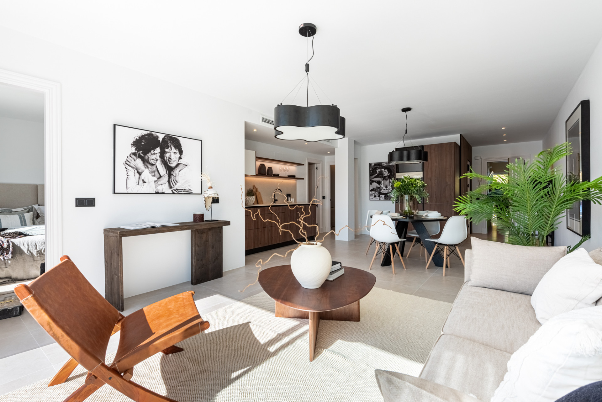 Open plan living area with light sofa, dining table, kitchen in background, and a view into a bedroom.