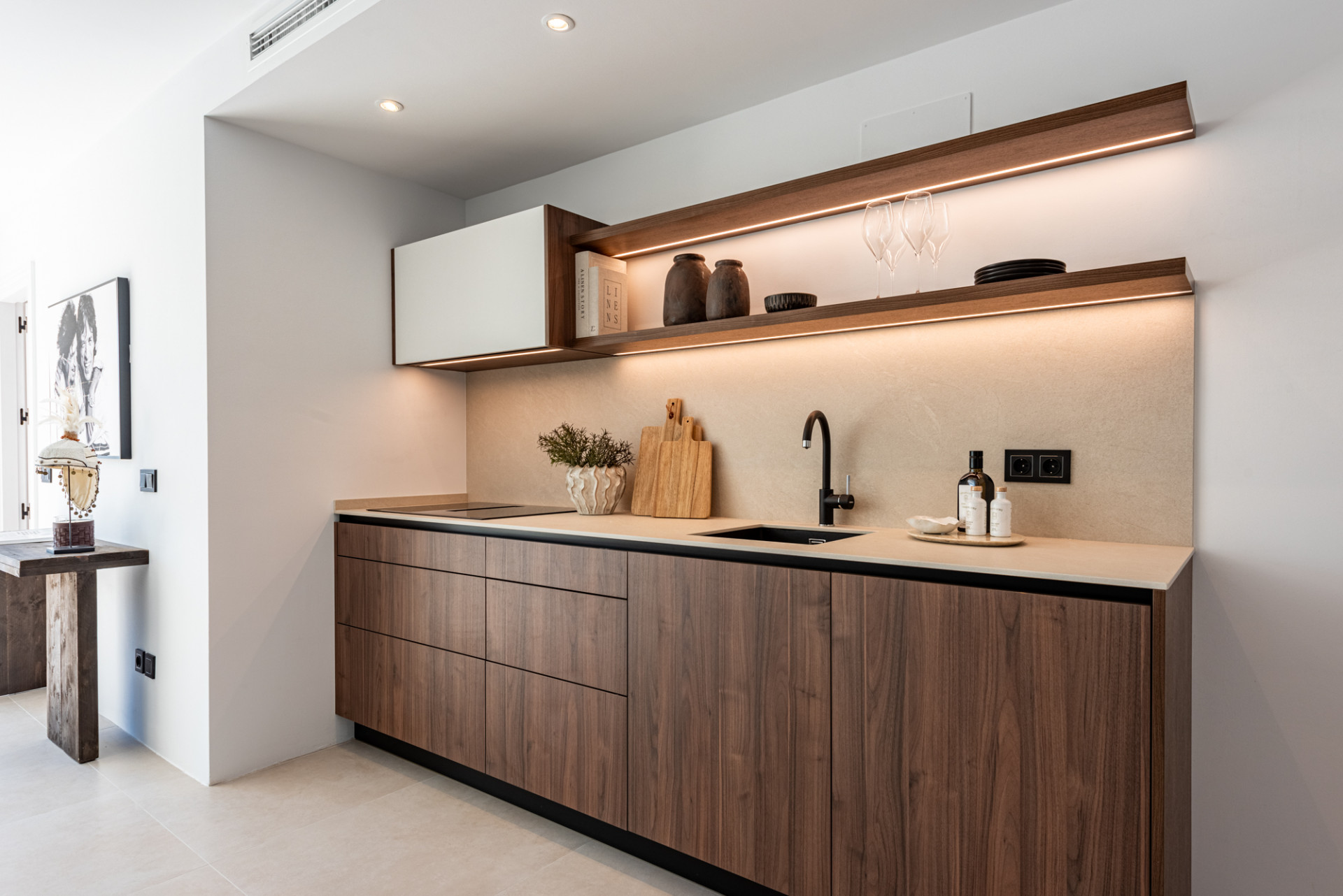 Modern kitchen with wood cabinets, light countertop, dark sink, and illuminated shelving above.