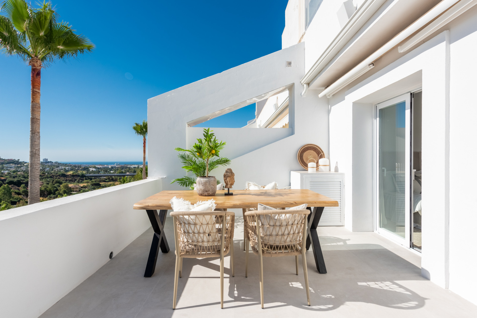 Outdoor terrace with a wooden dining table, four chairs, and a small plant, overlooking a city view.