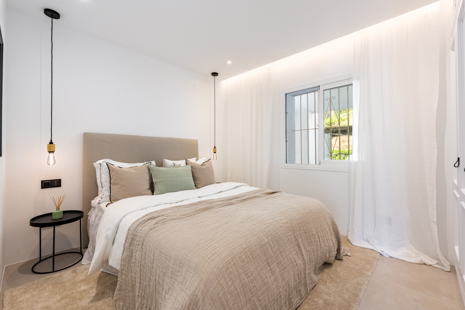 Bedroom with a large bed, light beige headboard, dark bedside table, and a window with white curtains.