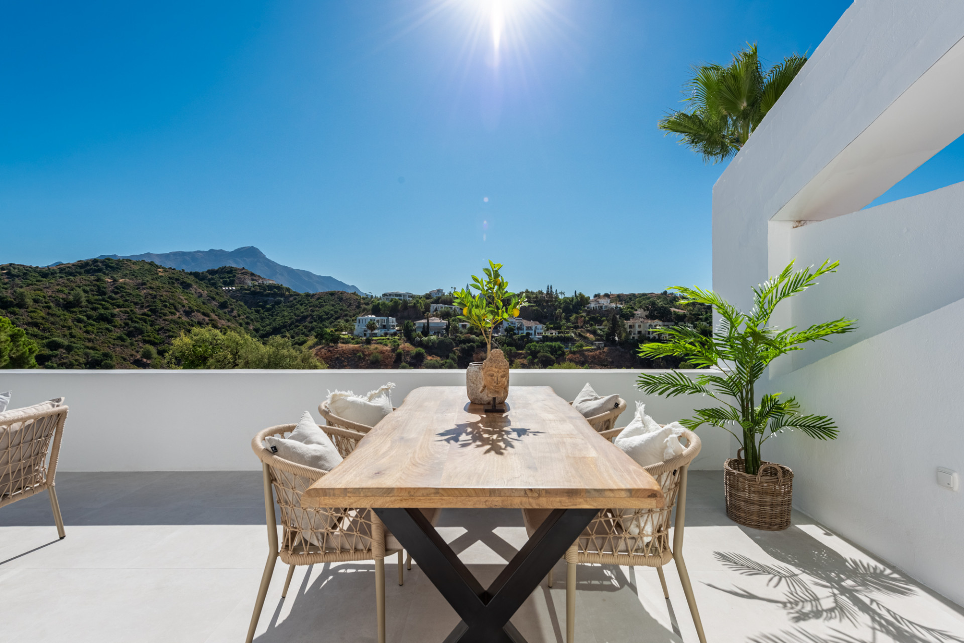 Sunny outdoor dining terrace with a large wooden table and six chairs, facing a green mountain landscape.