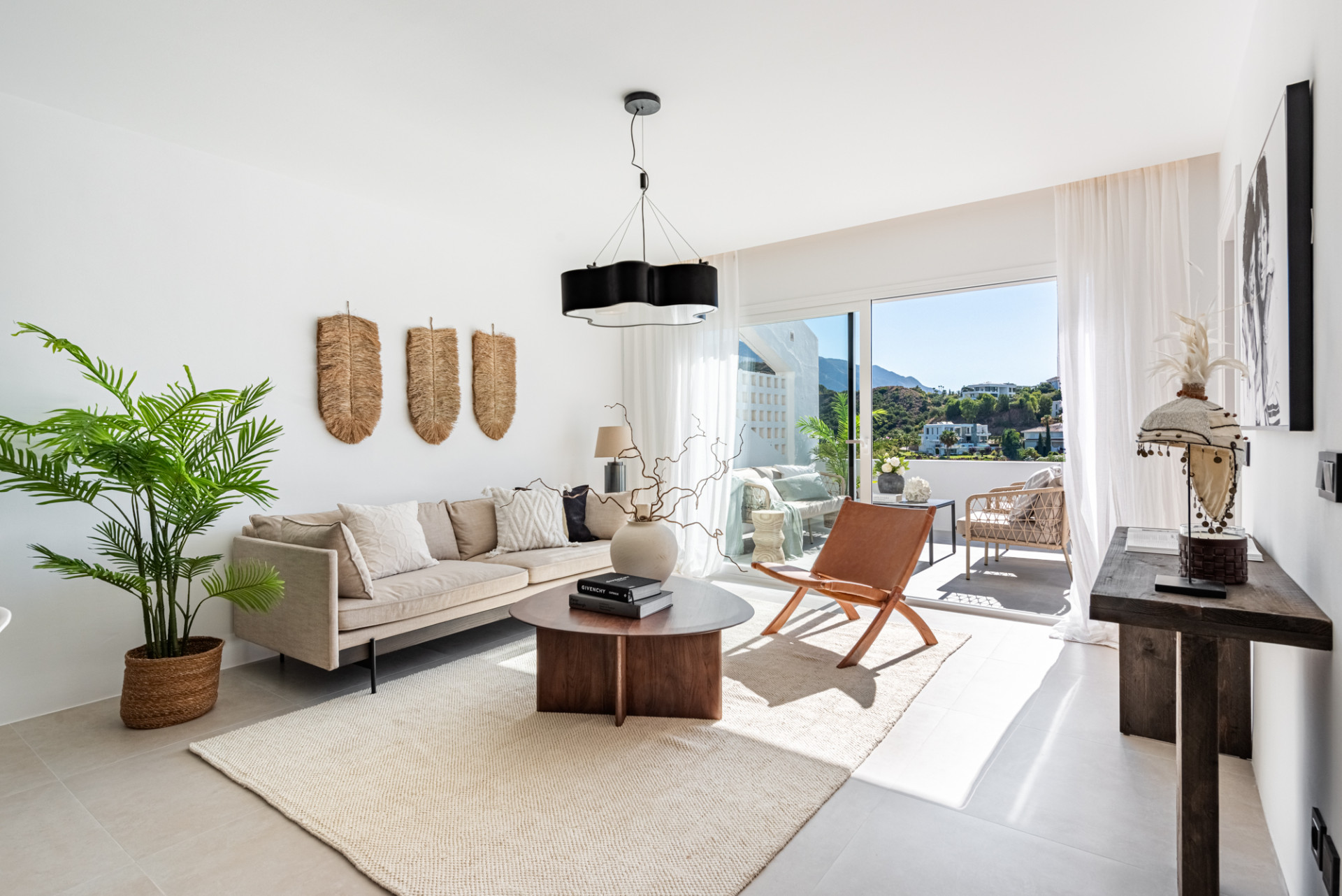 Bright living room with a cream sofa, a round coffee table, and a leather chair, opening to a balcony.