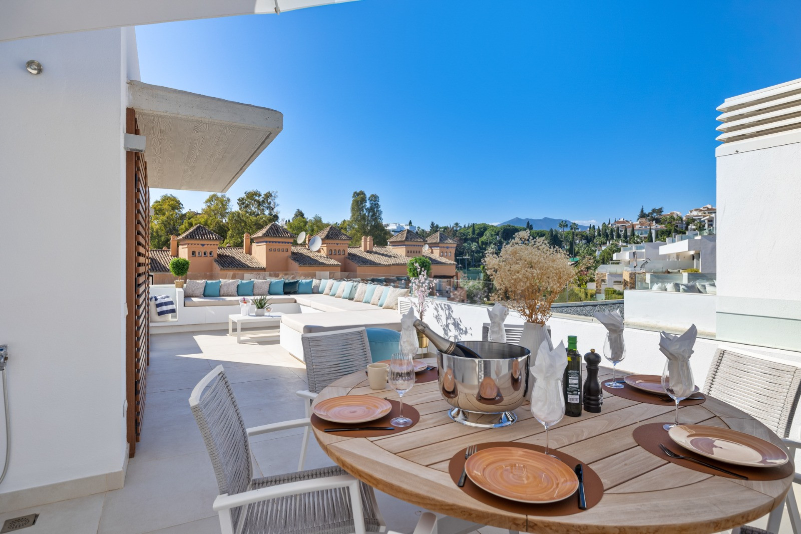 Covered terrace with a wooden dining table, plates arranged, and a view of surrounding architecture.